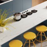 A white kitchen island of Snowdrift porcelain with delicate veining, yellow stools, and blue cabinets.